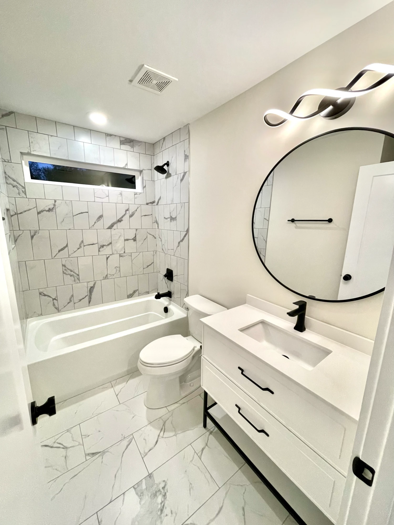 A bathroom in a new M3G home with marble-look tile, a tub, a white vanity, black fixtures and a round mirror