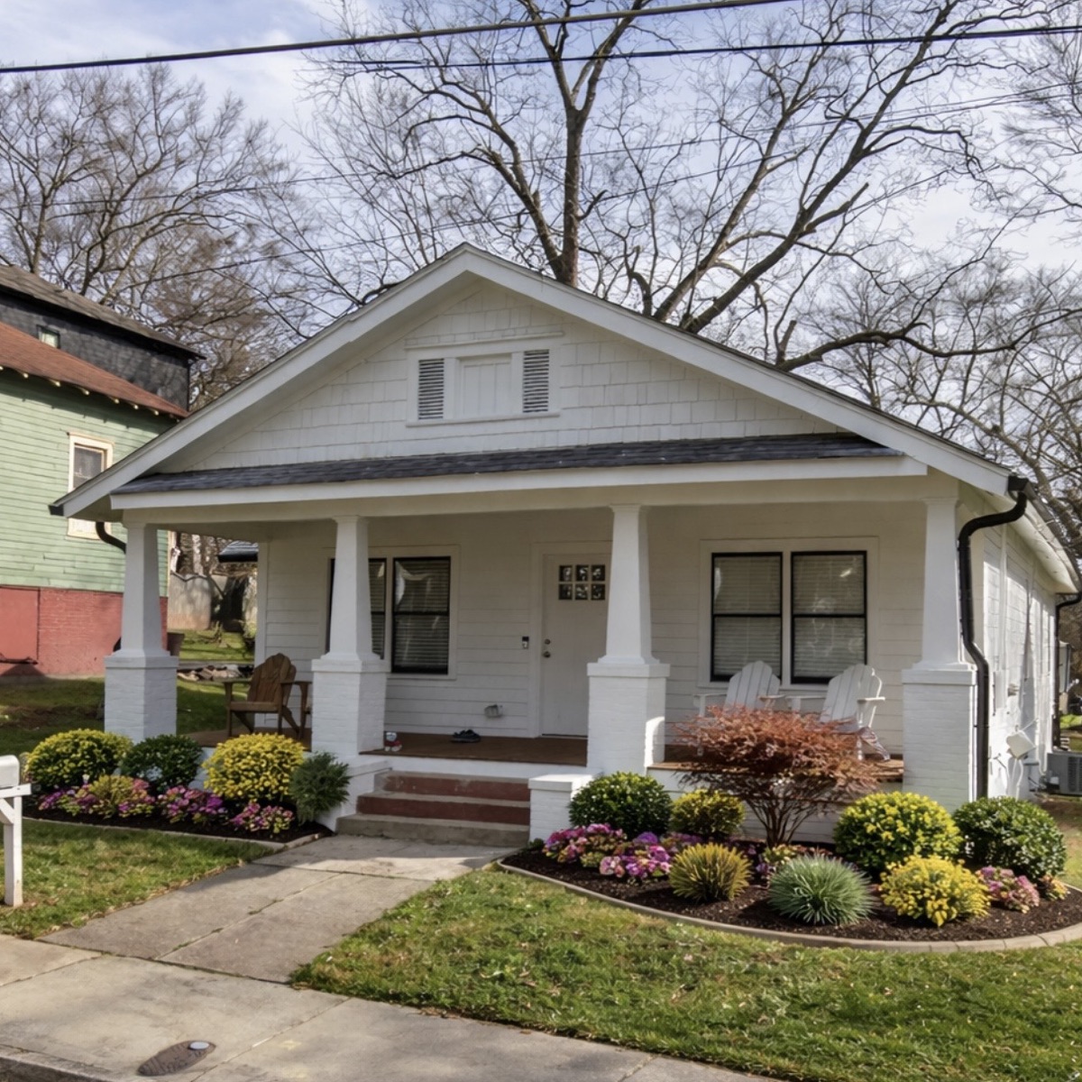 After: the same white bungalow with curved mulched beds full of shrubs and flowers along the porch and walk