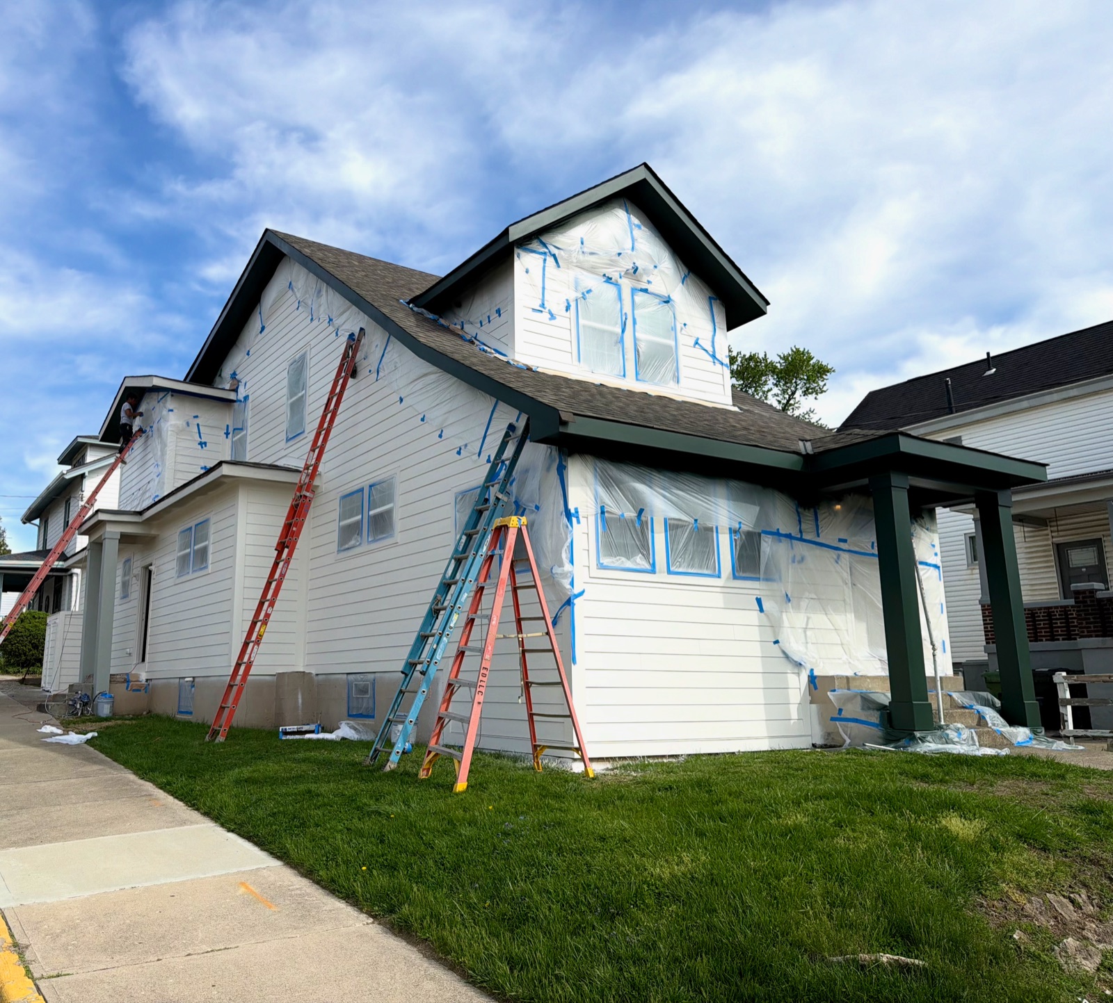 An M3G crew painting a white house, with windows and trim masked in plastic and blue tape and ladders against the siding