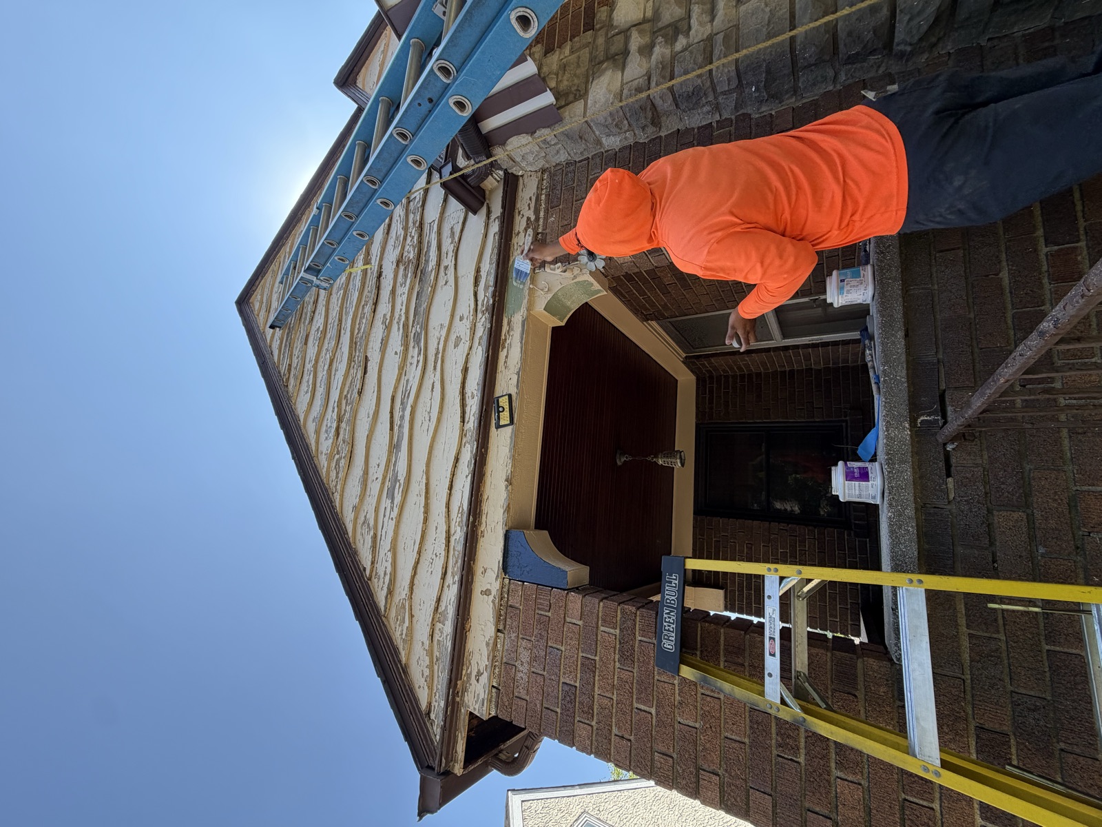 An M3G painter in an orange hoodie prepping the trim under a brick bungalow's gable