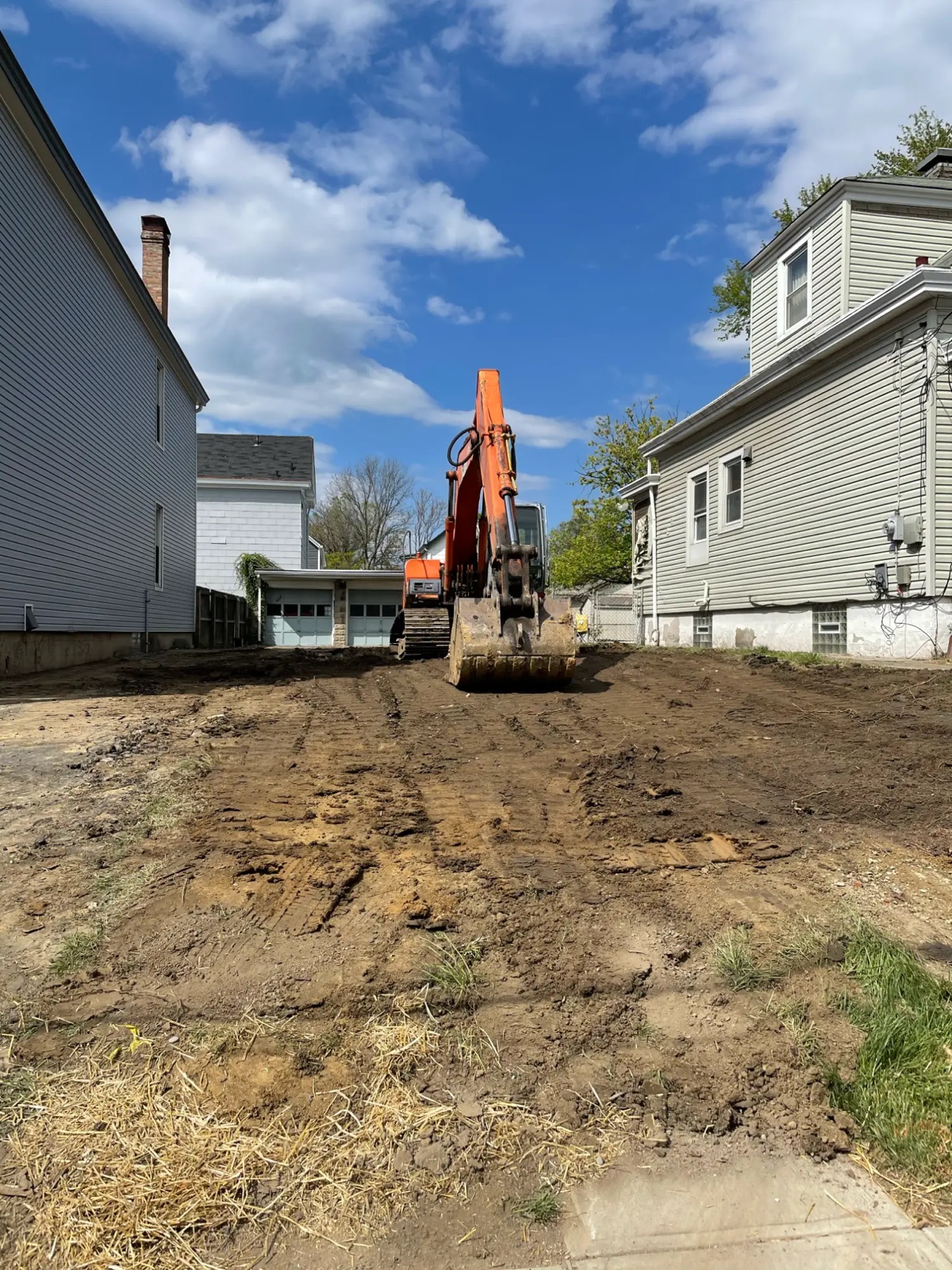 The cleared and graded lot with the excavator parked between the neighboring houses