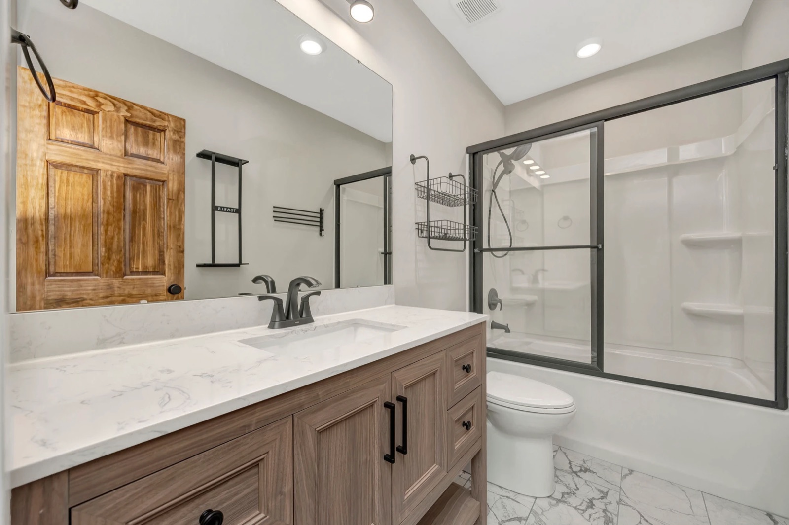 A bathroom with a wood vanity, marble-look counter, black fixtures and a tub with glass doors