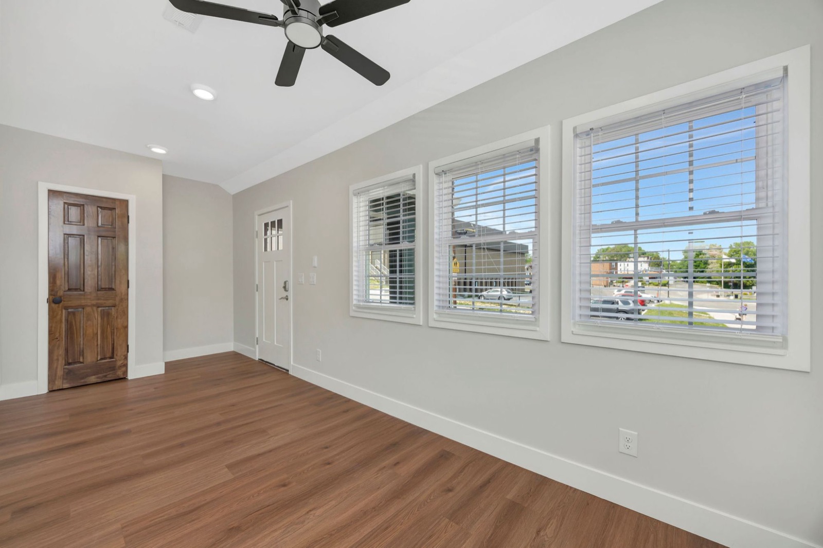 A bright front room with three large windows, a ceiling fan and a stained six-panel door